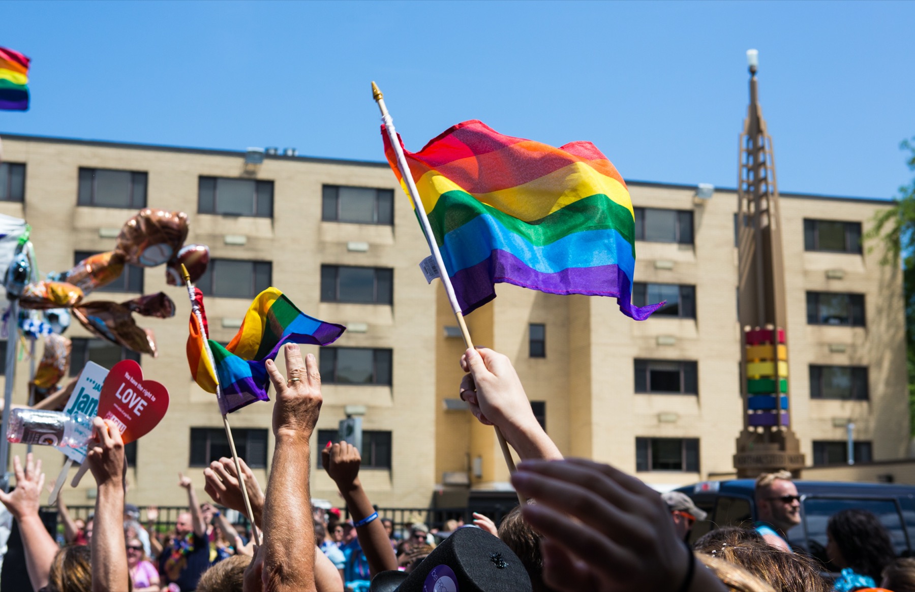 Rainbow flags wave above a joyful crowd at Chicago Pride Parade, with the iconic rainbow Legacy Walk pillars visible in the background.